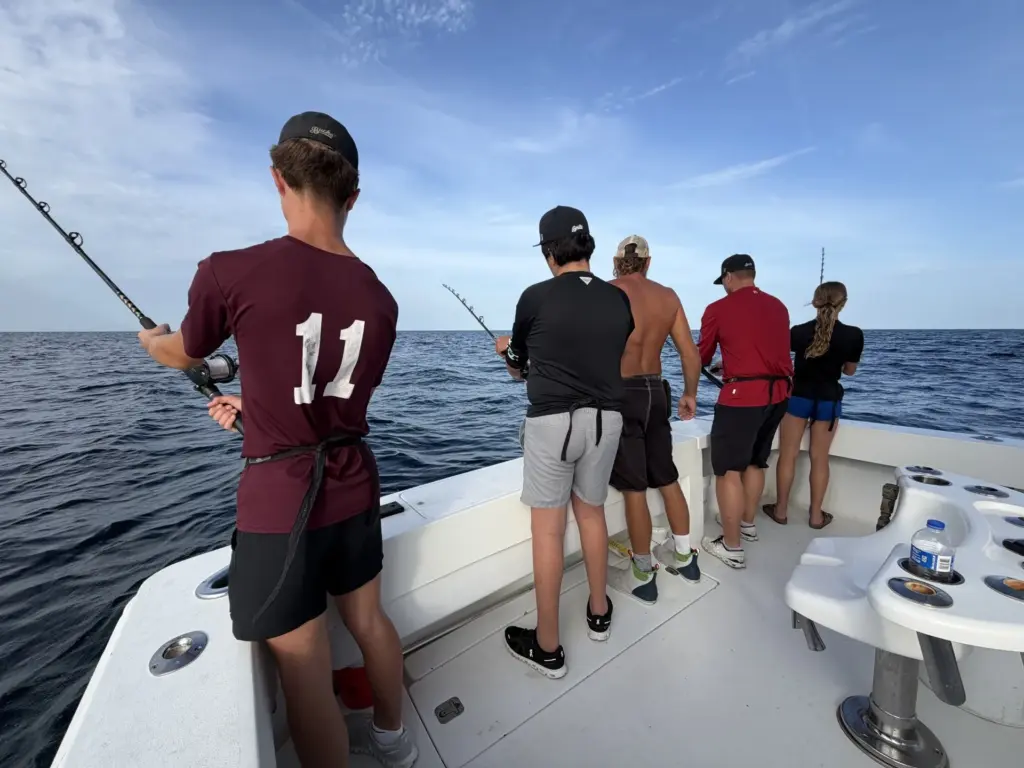 Five people stand along the stern of a charter boat holding fishing rods over open Gulf water under a clear sky.