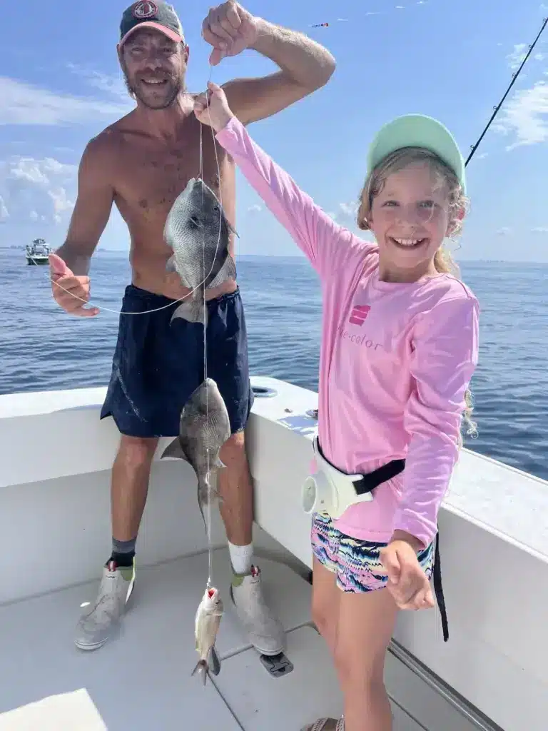 Smiling young girl in a pink shirt holds a fishing line with three fish aboard a boat while a man stands beside her.