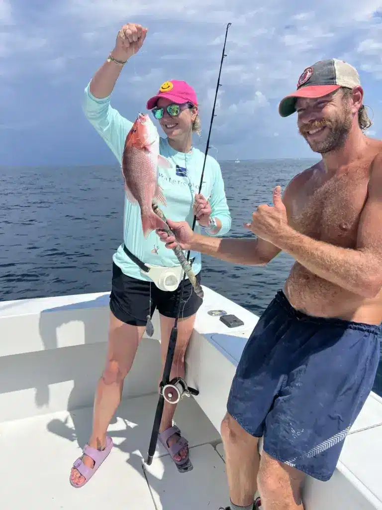Woman in a pink cap holds up a red snapper on a fishing rod while a man gives a thumbs up on a boat in the Gulf.