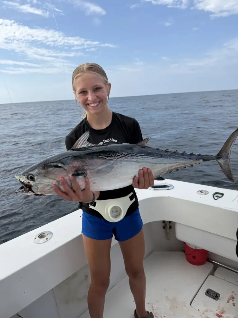 Smiling girl on a boat deck holding a large false albacore with both hands, open water and sky behind her.