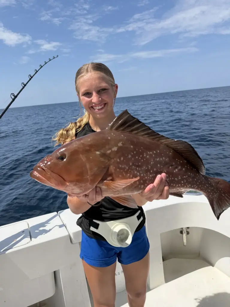 Smiling young woman holding a large red grouper on a charter fishing boat with open water behind her.