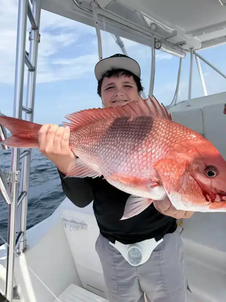Young angler in a black shirt and gray cap holds a large red snapper on a charter boat deck with the ocean behind.